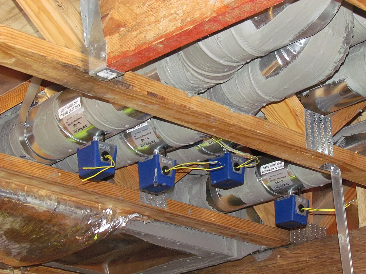 Metal supply ducts and flexible ducts running between floor joists in a basement ceiling, with small motorized dampers on each branch